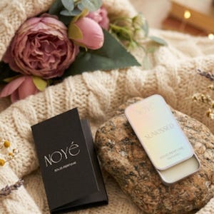 May include: A rectangular metal tin of NOYÉ Sunkissed solid perfume with an iridescent finish, resting on a textured stone next to a black folded card featuring the white text NOYÉ SOLID PERFUME. The tin is partially open, revealing the solid wax.