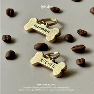 Op de afbeelding: Twee goudkleurige hondenpenningen in de vorm van een bot met de namen "Norman" en "Archie" in zwart gegraveerd. De penningen zitten aan een gouden ring. Koffiebonen zijn verspreid rond de penningen op een neutrale achtergrond. De tekst "MINIMAL ROAST" staat onderaan.