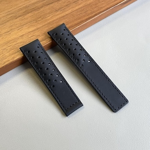 Può includere: Due cinturini neri per orologio con un design perforato. I cinturini sono realizzati in un materiale liscio e presentano fori distanziati uniformemente. I cinturini sono mostrati su una superficie grigio chiaro con uno sfondo in legno.