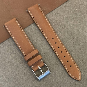 Peut inclure: Deux bracelets de montre en cuir marron clair avec des coutures blanches. L'un est doté d'une boucle argentée et d'une boucle. L'autre comporte des trous de réglage. Sur une surface marron.