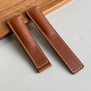 Könnte beinhalten: Zwei braune Uhrenarmbandsegmente aus Leder mit glatter, matter Oberfläche und markanten weißen Kontrastnähten entlang der Kanten. Die Armbänder haben eine rechteckige Form mit leicht abgerundeten Ecken und ein einheitliches Design.