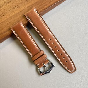 Peut inclure: Un bracelet de montre en cuir marron clair composé de deux parties avec des surpiqûres blanches contrastantes sur les bords. Il comprend une boucle ardillon en métal argenté poli, deux passants en cuir et sept trous de réglage.