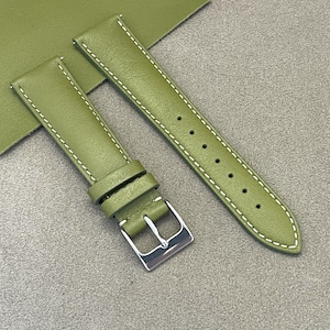 Peut inclure: Bracelet de montre en cuir vert olive avec surpiqûres blanches. Le bracelet est doté d'une boucle argentée et de plusieurs trous de réglage. Le bracelet est présenté sur une surface grise.