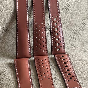 Può includere: Sei cinturini per orologi in pelle marrone con cuciture bianche. Tre cinturini hanno un design pieno e tre hanno un design traforato. I cinturini sono disposti su una superficie strutturata.