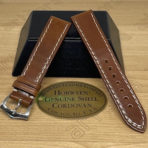 May include: Two brown leather watch straps with white stitching and a silver buckle. The straps are displayed on a wooden surface with a black box in the background. A brass plate reads "HORWEEN LEATHER COMPANY GENUINE SHELL CORDOVAN CHICAGO, ILL. U.S.A."