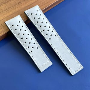 Peut inclure: Une paire de bracelets de montre gris clair présentant un motif perforé avec de petits trous circulaires disposés en grille. Les bracelets sont dotés de coutures blanches visibles sur les bords, offrant une apparence nette et structurée.