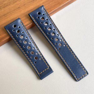 Può includere: Una coppia di cinturini per orologio in pelle blu navy caratterizzati da un design traforato con fori circolari di varie dimensioni e cuciture arancioni a contrasto lungo i bordi. I cinturini sono rettangolari con finitura liscia.