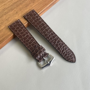 Könnte beinhalten: Ein Paar braune Uhrenarmbänder mit Krokodilleder-Muster. Die Armbänder sind aus Leder gefertigt und haben eine silberfarbene Schnalle. Die Uhrenarmbänder werden auf einer hellgrauen Oberfläche präsentiert.