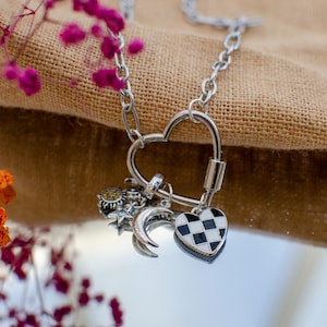 May include: A silver-toned necklace with a heart-shaped clasp and multiple charms. Charms include a checkered heart, a crescent moon, and a sunburst design. The necklace is displayed against a neutral fabric background with pink and orange floral accents.