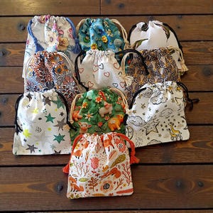 Peut inclure: Plusieurs sacs à cordon avec divers motifs, notamment floraux, cœurs, étoiles et animaux. Les sacs en tissu ont des cordons de serrage de différentes couleurs. Les sacs sont disposés sur une surface en bois.