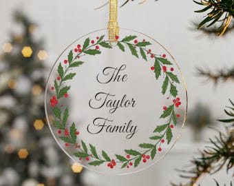 May include: A clear, round Christmas ornament with a gold ribbon for hanging. The ornament features a wreath of green leaves and red berries around the edge. The text "The Taylor Family" is written in elegant script in the center.