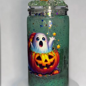 May include: A clear glass tumbler with a green glittery liquid inside, decorated with a cartoon ghost emerging from a carved pumpkin. The lid is adorned with a small orange pumpkin and a white ghost figurine.