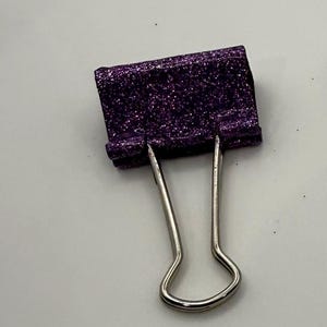 May include: A purple glitter binder clip with silver metal handles. The clip is a vibrant shade of purple and covered in sparkling glitter. The metal handles are curved and extend downwards.