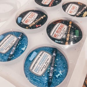 May include: A clear tray holds several resin coasters with school-themed designs. Each coaster features a composition book label, a crayon, and other small school supplies. The coasters have either a blue or black glitter background.