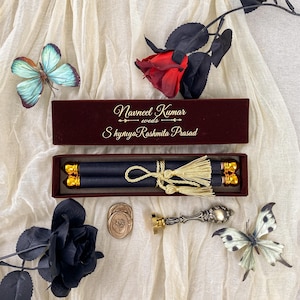 Peut inclure: Une boîte rectangulaire marron foncé avec le texte doré Navneel Kumar weds ShyniyaRashmita Prasad, contenant un parchemin bleu foncé noué de pompons dorés, un sceau en métal et deux sceaux de cire, entourés de deux papillons et deux roses.