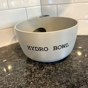 Op de afbeelding: Een witte, ronde kom met de woorden "HYDRO BOWL" in zwarte letters. De kom heeft een gat in het midden en staat op een donker aanrecht. De achtergrond is voorzien van witte metro tegels.