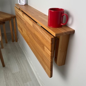 May include: A wooden folding table, mounted on a white wall, with a red mug and a small white alarm clock on top. The table is partially folded down, showing its construction. The table is made of light-colored wood.