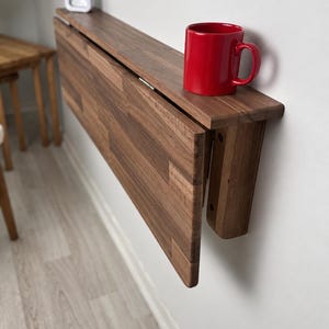 May include: A wall-mounted, foldable wooden table made of dark brown wood with a visible grain pattern. The table is in a folded, vertical position against a white wall. A bright red ceramic mug sits on the top edge of the folded table.