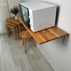 May include: A white microwave oven with a black door is mounted on a wooden shelf attached to a white wall. A small wooden stool and a set of nesting tables are nearby. The microwave has two control knobs.