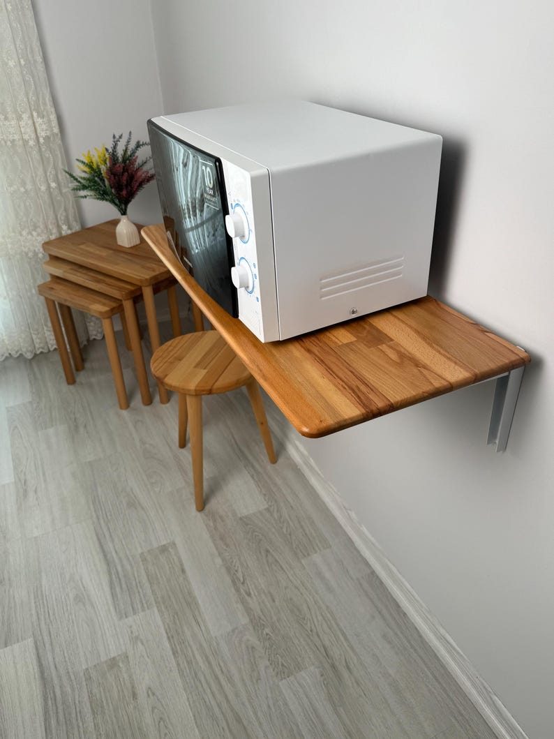 May include: A white microwave oven with a black door is mounted on a wooden shelf attached to a white wall. A small wooden stool and a set of nesting tables are nearby. The microwave has two control knobs.