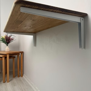 May include: A wall-mounted wooden desk with a light brown finish and a rounded edge. The desk is supported by white metal brackets. The wood grain is visible. The desk is approximately 36 inches long.