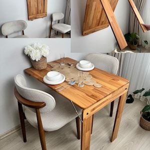 Könnte beinhalten: Eine hölzerne, an der Wand befestigte Klapptisch, im geschlossenen und geöffneten Zustand gezeigt. Der Tisch ist im geöffneten Zustand rechteckig, mit zwei Stühlen und einer Tischdekoration. Der Tisch ist aus hellem Holz gefertigt. Der geschlossene Tisch ist eine rechteckige Platte.