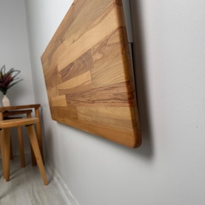 May include: A wall-mounted, rectangular wooden table with a natural wood grain finish. The table is attached to a white bracket on a light gray wall. The table's edge is rounded.