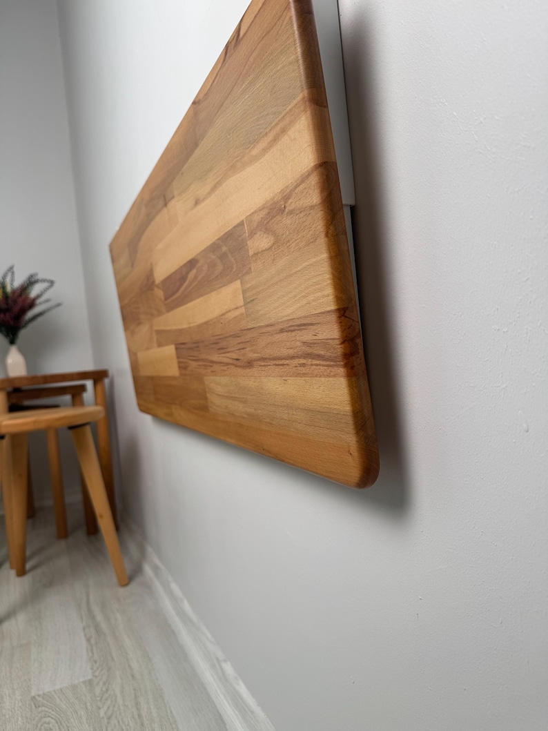 May include: A wall-mounted, rectangular wooden table with a natural wood grain finish. The table is attached to a white bracket on a light gray wall. The table's edge is rounded.