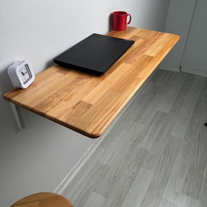 May include: A wall-mounted wooden desk with a black laptop, a red mug, and a white alarm clock. The desk is made of light-colored wood and is supported by a white bracket. The room has light gray walls and flooring.