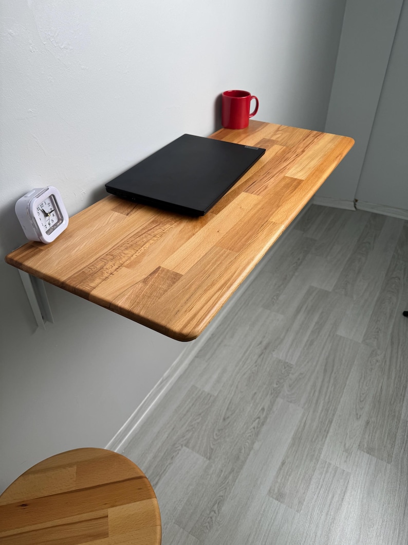 May include: A wall-mounted wooden desk with a black laptop, a red mug, and a white alarm clock. The desk is made of light-colored wood and is supported by a white bracket. The room has light gray walls and flooring.