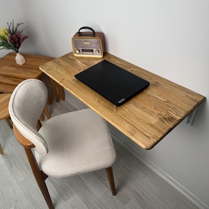 May include: A wooden wall-mounted desk with a black laptop and a vintage radio. A beige upholstered chair with wooden legs sits in front of the desk. The desk is attached to a white wall. A small wooden table and vase with flowers are in the background.