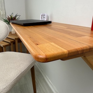May include: A wooden desk with a rounded edge is attached to a white wall. A black laptop and a small white alarm clock sit on the desk. A beige chair with wooden legs is in the foreground.