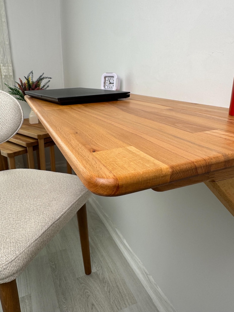 May include: A wooden desk with a rounded edge is attached to a white wall. A black laptop and a small white alarm clock sit on the desk. A beige chair with wooden legs is in the foreground.