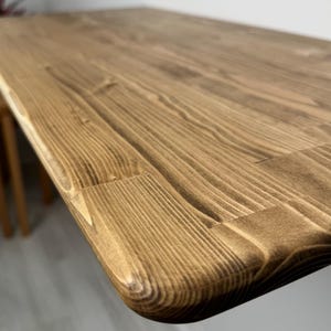May include: Close-up of a wooden table with a warm, brown stain. The table's surface showcases prominent wood grain and a rounded edge. The table appears to be made of solid wood, with a natural finish. The table is a piece of furniture.