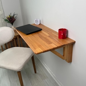 May include: A light-coloured wooden folding desk mounted on a white wall. A black laptop, a small white clock, and a red mug are on the desk. A beige chair with wooden legs is in the foreground. A vase with flowers is on a small table.
