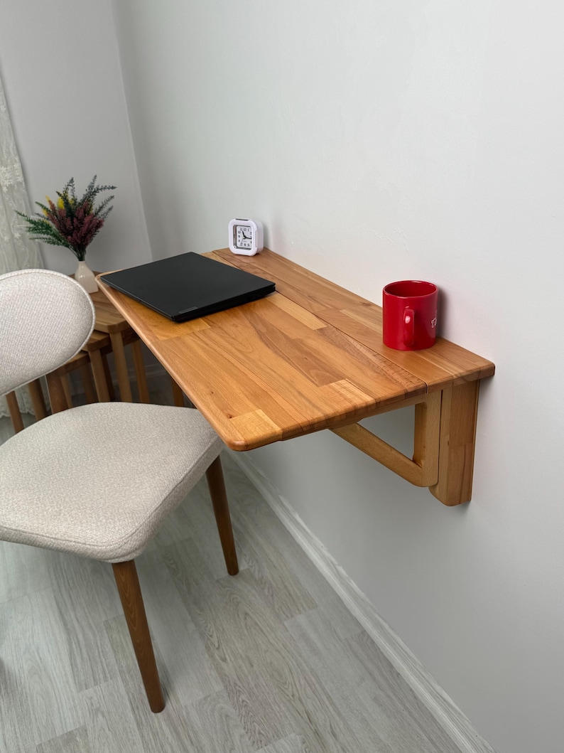 May include: A light-coloured wooden folding desk mounted on a white wall. A black laptop, a small white clock, and a red mug are on the desk. A beige chair with wooden legs is in the foreground. A vase with flowers is on a small table.