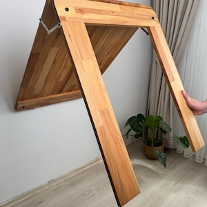 Könnte beinhalten: Ein hölzerner, wandmontierter Klapptisch in natürlicher Holzoptik. Der Tisch ist rechteckig mit abgerundeten Ecken und wird von zwei abgewinkelten Beinen getragen. Der Tisch ist mit Metallscharnieren an der Wand befestigt. Der Tisch ist teilweise heruntergeklappt.