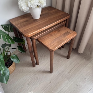 Puede incluir: Un juego de tres mesas nido de madera oscura. Las mesas tienen tapas rectangulares y esquinas redondeadas. Un jarrón blanco con flores blancas se encuentra sobre la mesa más grande. Las mesas están sobre un suelo de madera clara.