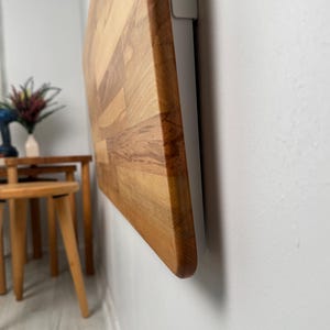 May include: A wooden, rectangular cutting board with a light brown and reddish-brown color scheme is mounted on a white wall. The board is made of multiple wood pieces joined together. The edge of the board is rounded.