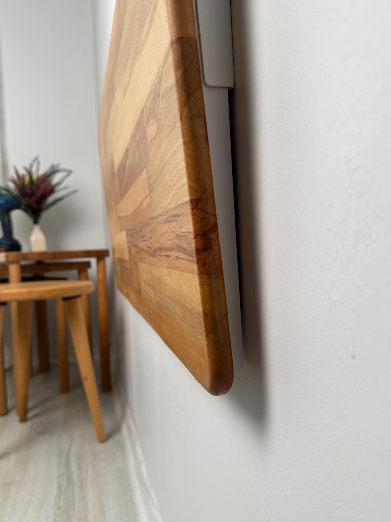 May include: A wooden, rectangular cutting board with a light brown and reddish-brown color scheme is mounted on a white wall. The board is made of multiple wood pieces joined together. The edge of the board is rounded.