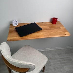 May include: A light-coloured wooden wall-mounted desk with a black laptop, a red mug, and a white alarm clock. A grey and wood-toned chair sits below the desk. The desk is attached to a white wall.