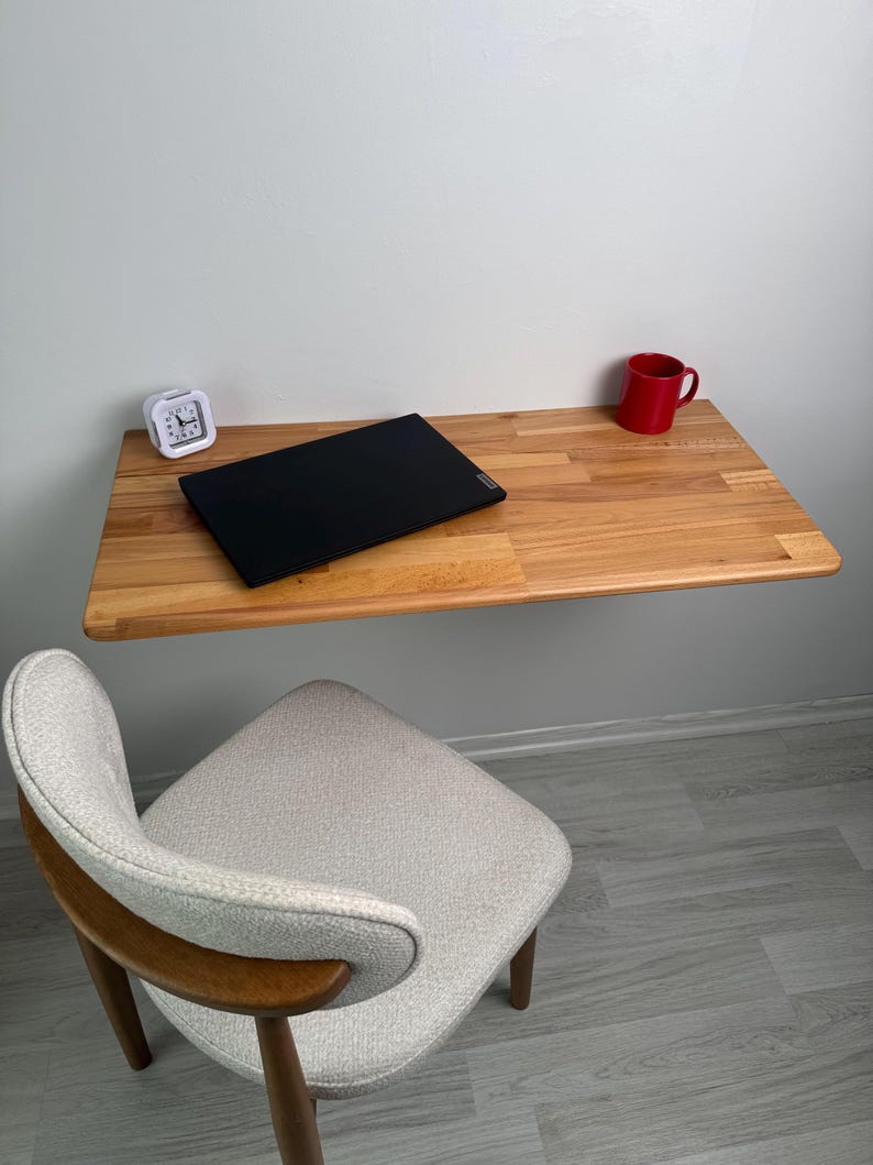 May include: A light-coloured wooden wall-mounted desk with a black laptop, a red mug, and a white alarm clock. A grey and wood-toned chair sits below the desk. The desk is attached to a white wall.