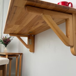 May include: A light-coloured wooden, wall-mounted folding table. The table is rectangular with rounded corners and supported by a wooden bracket. A red mug sits on the table's surface. The table is designed to save space.