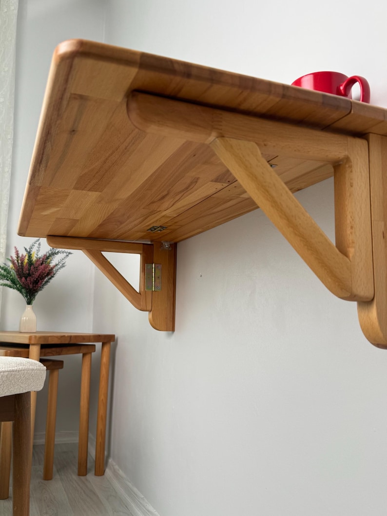 May include: A light-coloured wooden, wall-mounted folding table. The table is rectangular with rounded corners and supported by a wooden bracket. A red mug sits on the table's surface. The table is designed to save space.