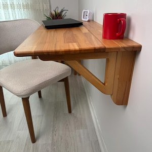 May include: A wooden, wall-mounted folding desk with a red mug on top. A laptop and small clock are also on the desk. A beige chair with wooden legs is in front of the desk. The desk is made of light-coloured wood.