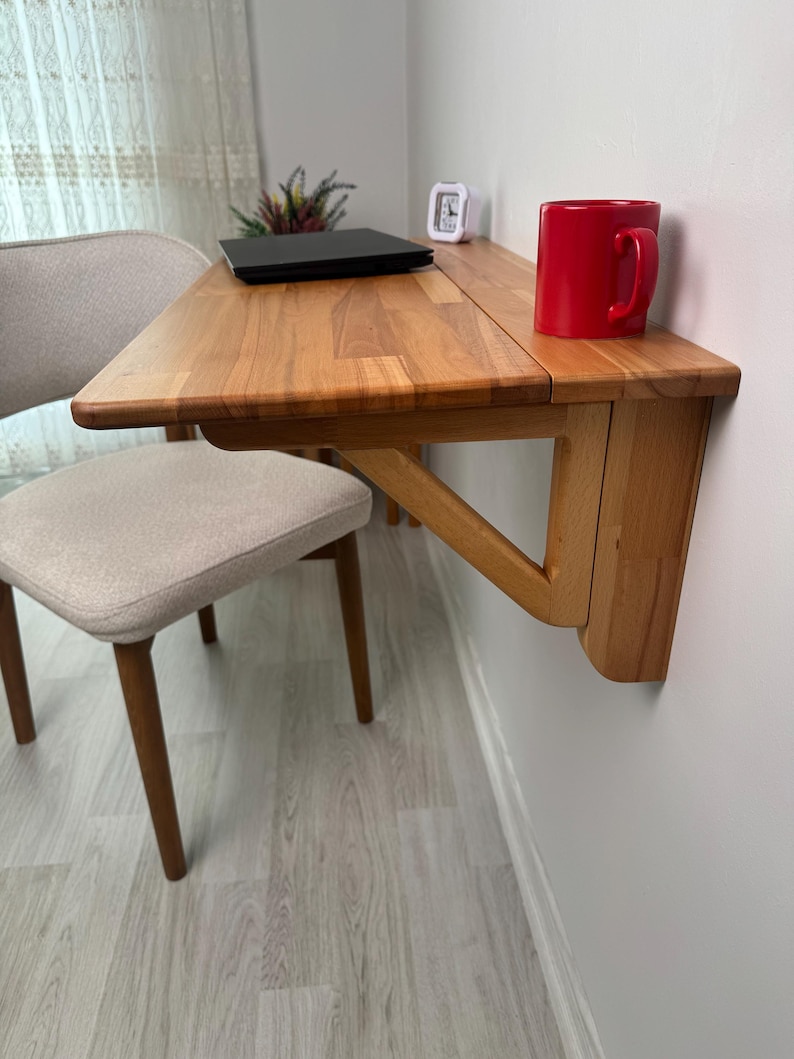 May include: A wooden, wall-mounted folding desk with a red mug on top. A laptop and small clock are also on the desk. A beige chair with wooden legs is in front of the desk. The desk is made of light-coloured wood.