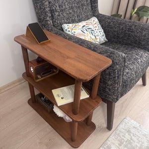 Op de afbeelding: Een houten bijzettafel met drie niveaus en afgeronde hoeken, afgewerkt in donkerbruin. De bovenste plank bevat een zwarte smartphone op een standaard, de middelste plank een vintage radio en een boek, en de onderste plank een klein mandje.