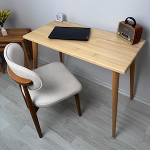 Puede incluir: Un escritorio de madera clara con un portátil negro, un pequeño reloj blanco y una radio de estilo vintage. Una silla tapizada beige con brazos y patas de madera está frente al escritorio. Una pequeña mesa auxiliar de madera con un jarrón de flores amarillas está a la izquierda.