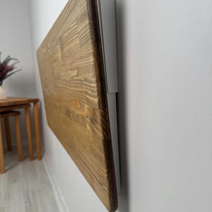 May include: A wall-mounted, folding wooden table with a natural wood grain finish. The table is rectangular and attached to a gray metal bracket. The wood has a warm, brown tone. The table is folded against a white wall.