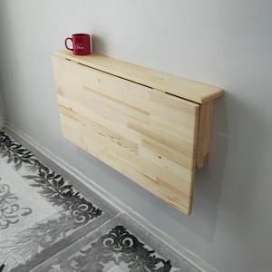 May include: A light-coloured, wall-mounted, folding table made of wood. A red mug with white text sits on the table's surface. The table is mounted on a light grey wall, above a patterned rug.